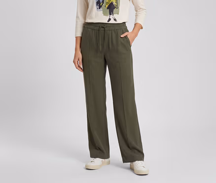 Personne portant un pantalon large vert olive avec cordon de serrage à la taille, des baskets blanches et un t-shirt clair à manches longues. Une main est dans la poche du pantalon.
