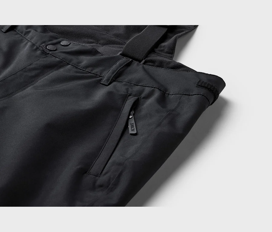 Gros plan d'un pantalon technique noir avec des boutons, une poche zippée avec tirette "ACTIVE" et une bretelle.