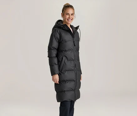 Femme souriante portant un long manteau doudoune noir à capuche et un pantalon foncé, debout devant un fond clair.