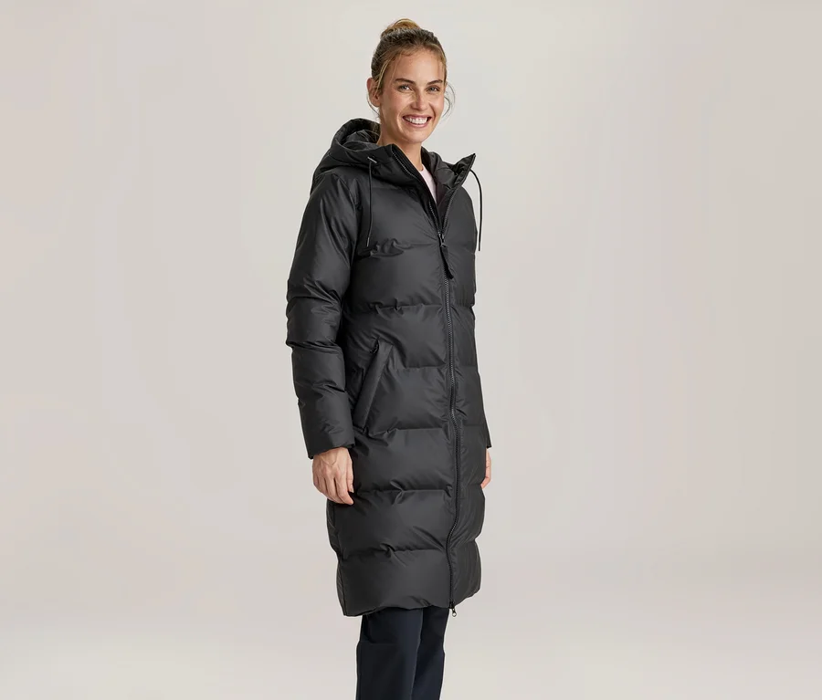 Femme souriante portant un long manteau doudoune noir à capuche et un pantalon foncé, debout devant un fond clair.