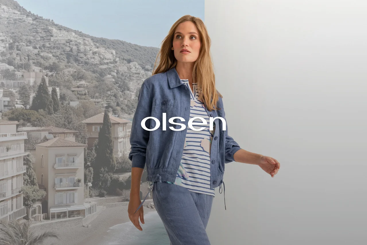 Femme en veste bleue, haut rayé et pantalon bleu, marchant devant un fond divisé entre une ville côtière et un mur gris, avec le texte 'olsen' superposé.