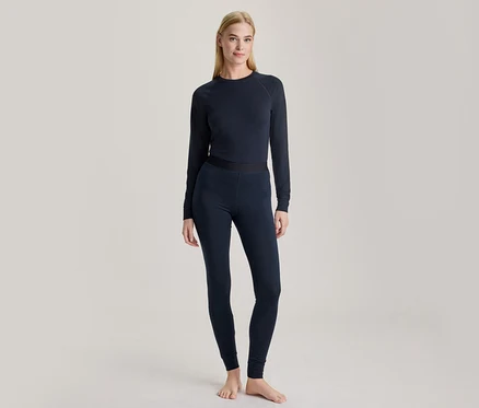 Une femme blonde est debout pieds nus, vêtue d'un haut à manches longues bleu foncé et d'un legging assorti.