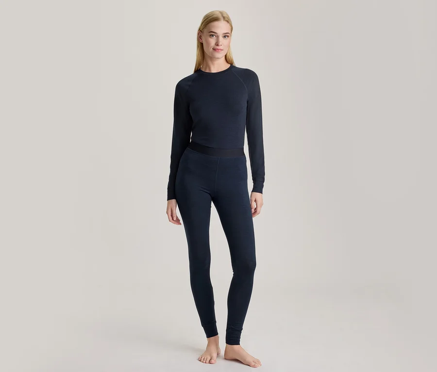 Une femme blonde est debout pieds nus, vêtue d'un haut à manches longues bleu foncé et d'un legging assorti.