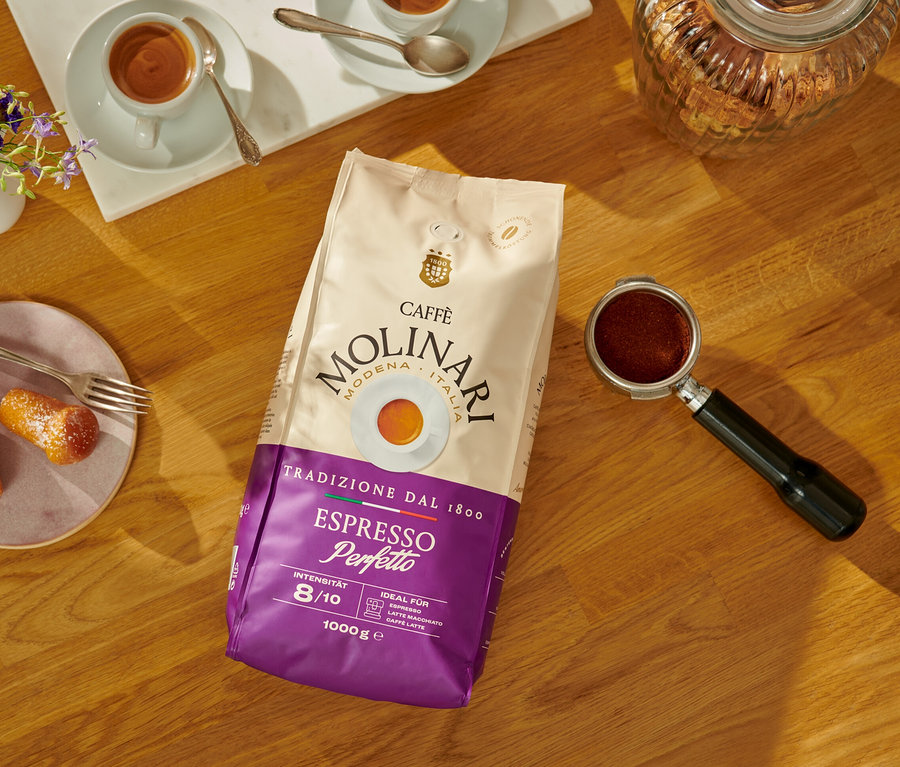 Sur une table en bois se trouvent un paquet de café en grains Caffè Molinari Espresso Perfetto, une tasse d'espresso et des pâtisseries.