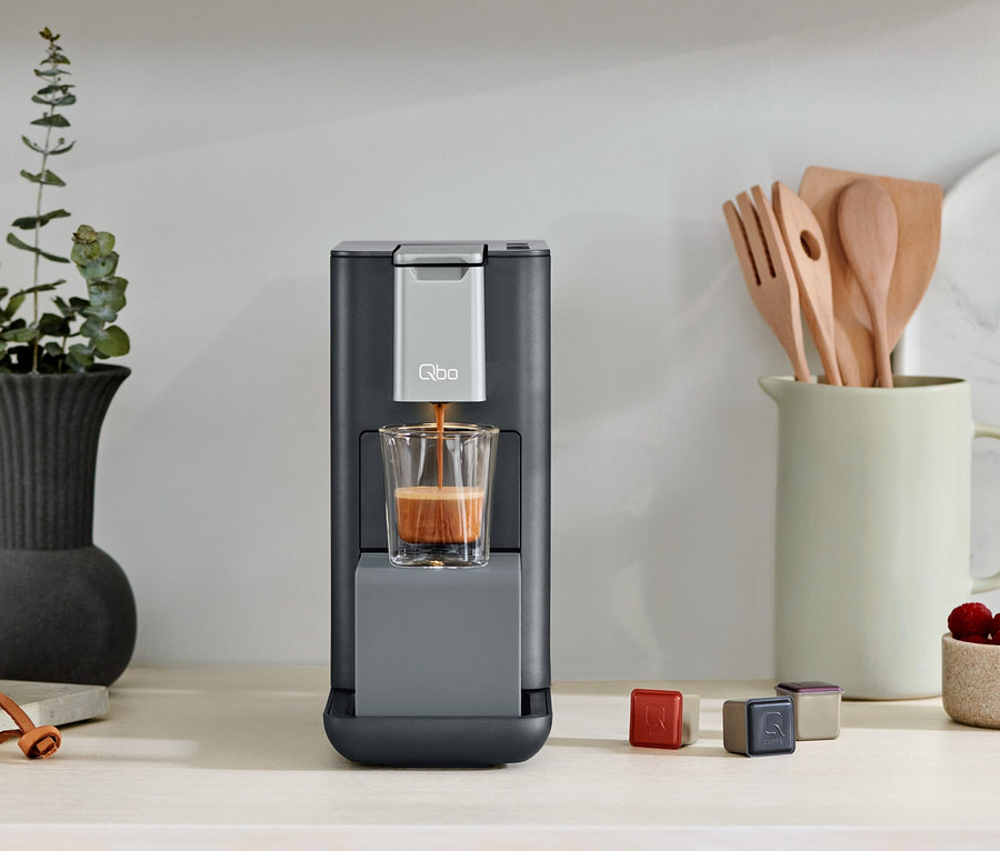 Une machine à café Qbo ESSENTIAL Dark Stone verse du café dans un verre. Des capsules sont à côté de la machine à café.