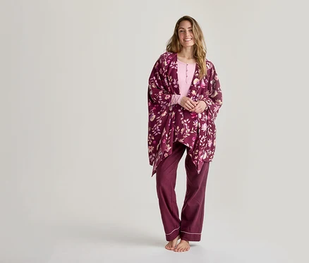 Femme souriante portant un haut rose pâle à manches longues, un pantalon de pyjama rouge foncé avec un liseré blanc et un kimono floral rouge foncé, debout pieds nus devant un fond blanc.