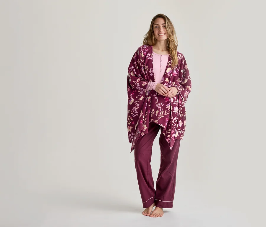 Femme souriante portant un haut rose pâle à manches longues, un pantalon de pyjama rouge foncé avec un liseré blanc et un kimono floral rouge foncé, debout pieds nus devant un fond blanc.