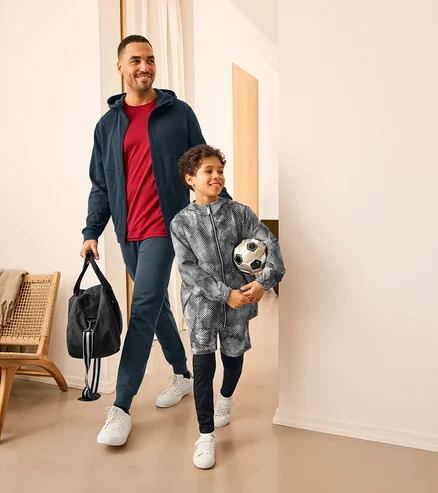 Un homme souriant et un garçon marchent ensemble. L'homme porte un sweat à capuche bleu foncé, un t-shirt rouge, un pantalon de jogging bleu foncé et des baskets blanches, et tient un sac de sport noir. Le garçon porte une veste à motifs noirs et blancs, un short assorti, des leggings foncés et des baskets blanches, et tient un ballon de football.