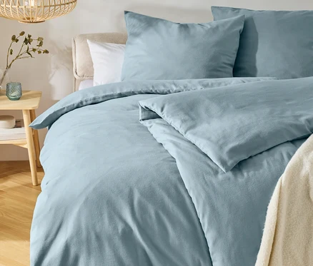 Lit avec une parure de lit bleu clair et deux oreillers, une tête de lit claire et un jeté de lit crème. À gauche, une table de chevet en bois avec un vase et un photophore, au-dessus pend une lampe en bois. Un tapis rond est posé sur le parquet.