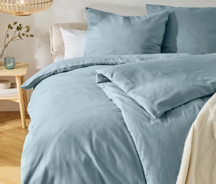 Lit avec une parure de lit bleu clair et deux oreillers, une tête de lit claire et un jeté de lit crème. À gauche, une table de chevet en bois avec un vase et un photophore, au-dessus pend une lampe en bois. Un tapis rond est posé sur le parquet.
