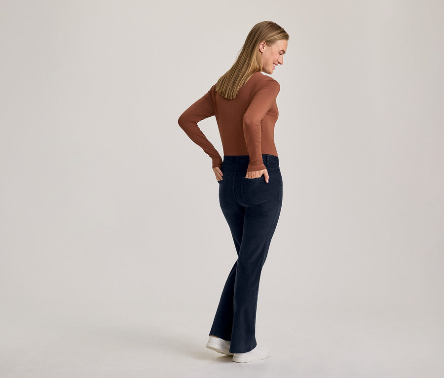 Femme de dos, légèrement tournée, portant un haut marron à manches longues et un pantalon évasé bleu foncé, les mains dans les poches et souriant légèrement.