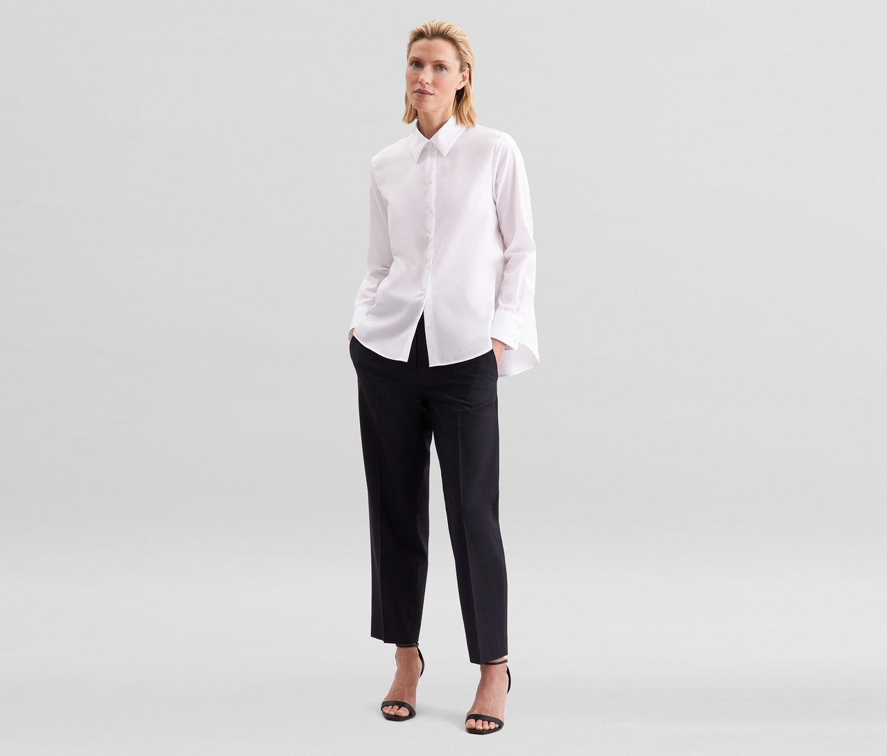 Femme blonde debout, portant un chemisier sans repassage Seidensticker blanc, un pantalon noir raccourci et des sandales noires à talons hauts.