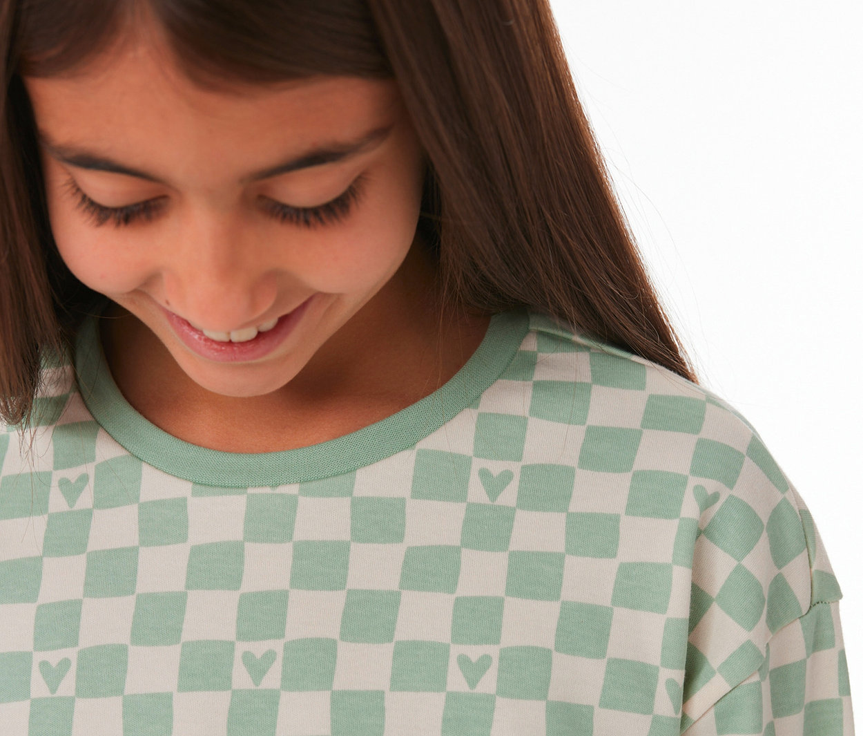 Jeune fille souriante regardant vers le bas, portant un haut de pyjama Interlock SCHIESSER à carreaux vert clair avec des cœurs.