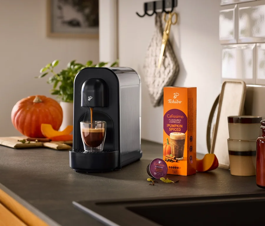 Machine à café noire Cafissimo now préparant un espresso dans un verre, à côté d'un paquet de café Cafissimo Pumpkin Spiced et d'une capsule, entourés de citrouille et d'épices sur un comptoir de cuisine.