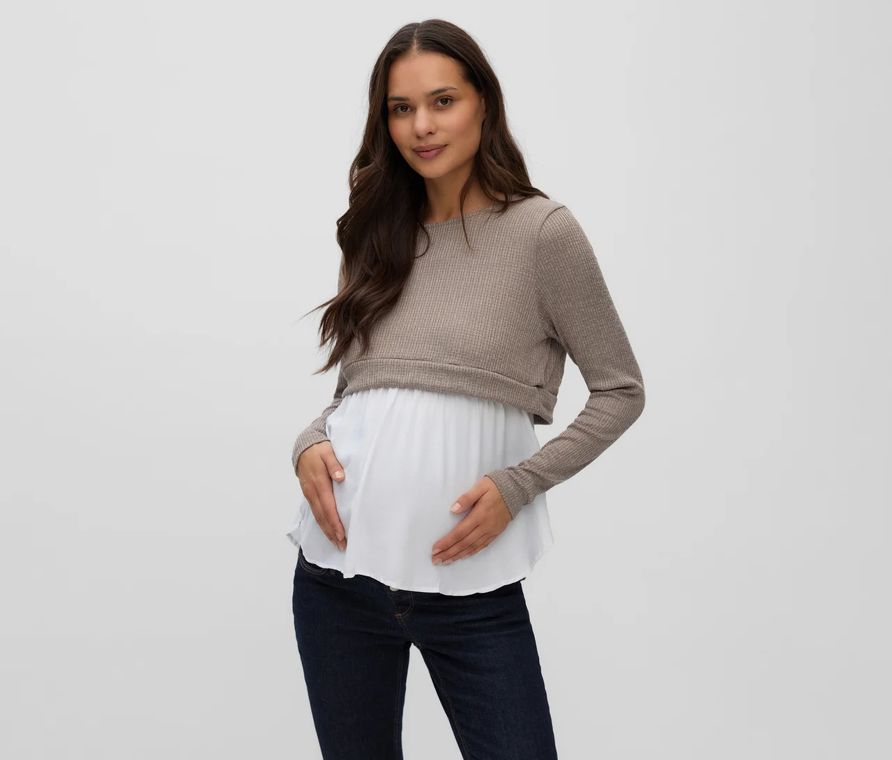 Une femme enceinte porte un haut mixte MAMALICIOUS « MELONI JUNE » avec un haut côtelé beige sur une couche blanche, et un jean bleu foncé. Elle tient doucement son ventre de femme enceinte.