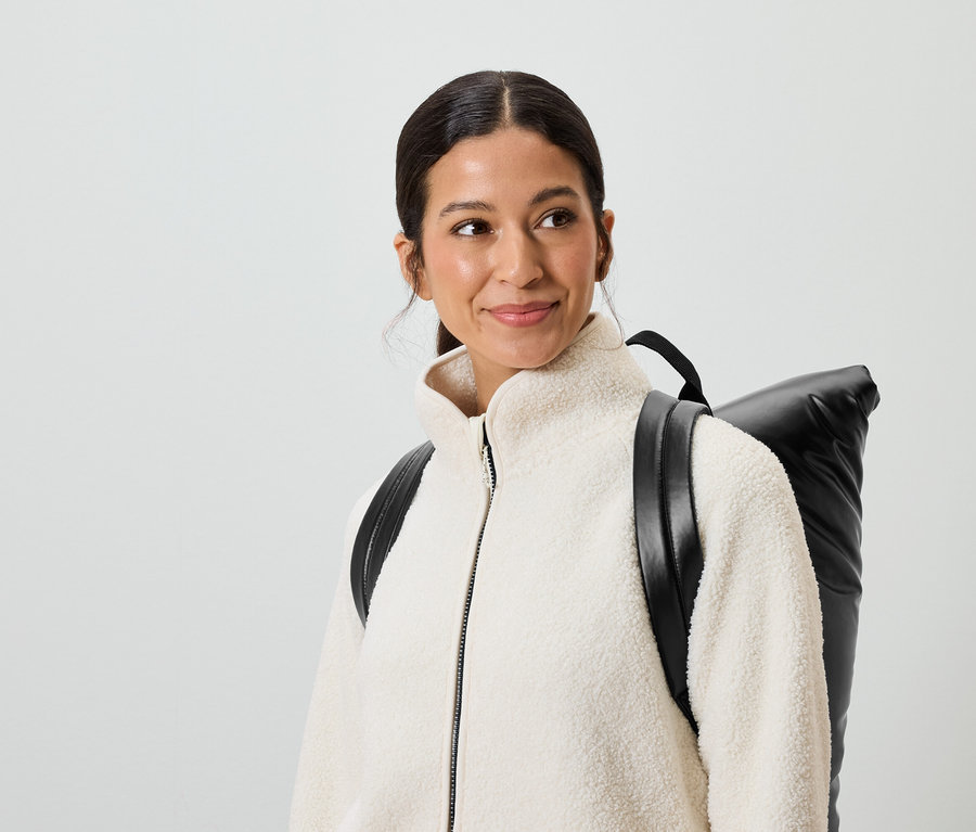 Une femme souriante aux cheveux bruns et aux yeux marron porte une veste crème et un sac à dos noir.