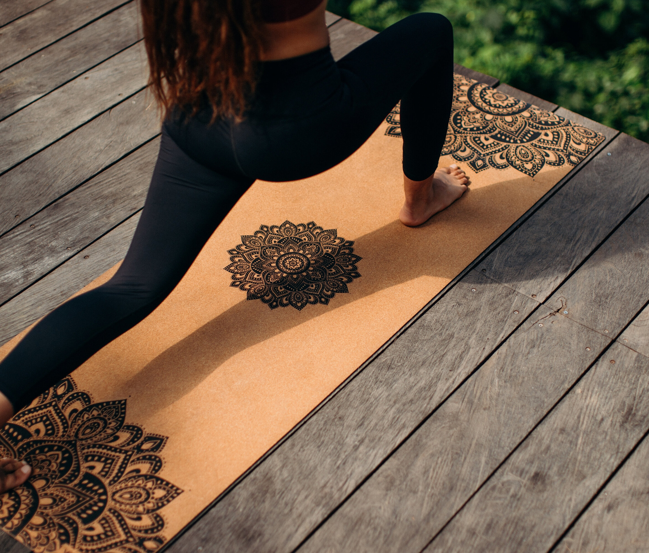 Une femme en leggings noirs fait la posture du guerrier sur un tapis de yoga beige avec des motifs de mandala sur un plancher en bois.