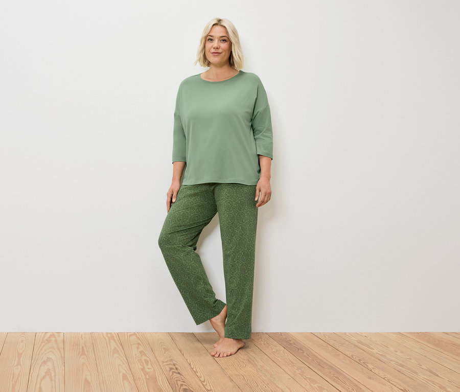Une femme blonde en pyjama vert se tient sur un plancher en bois devant un mur blanc.