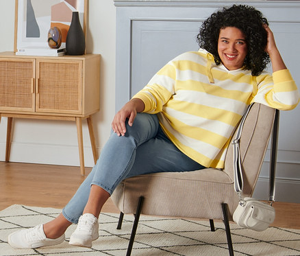 Une femme est assise dans un fauteuil et porte un sweat-shirt rayé à capuche jaune-crème, un tregging, des baskets en cuir velours et un sac porté épaule.