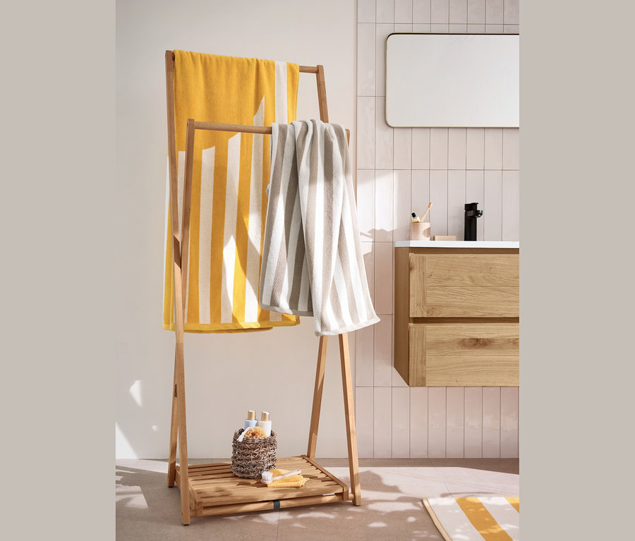 Un très grand linge de bain en jacquard jaune et blanc à rayures et 2 linges de toilette beige et blanc à rayures sont suspendus à un portant à vêtements pliable. En dessous se trouvent 2 paniers de rangement.