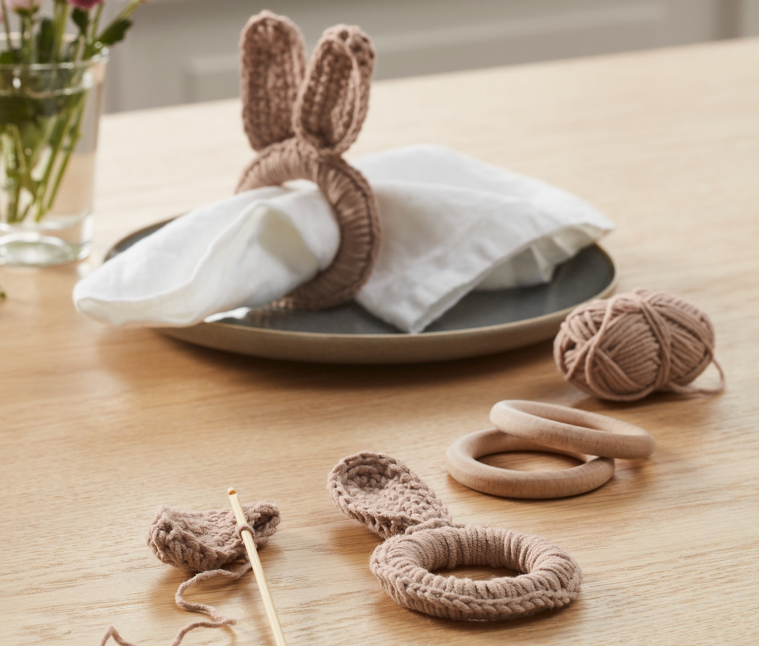 Sur une table se trouve un kit crochet DIY « ronds de serviette ». Un rond fini avec des oreilles de lapin est posé sur une serviette dans une assiette. À côté se trouvent un ouvrage au crochet en cours, une pelote de laine et des anneaux en bois.