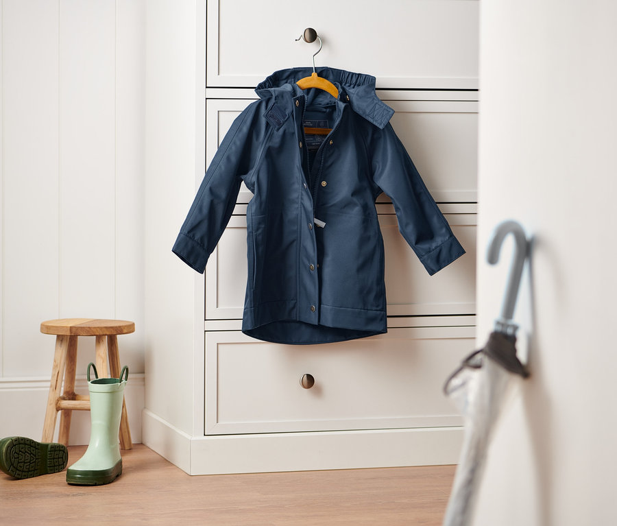 Une veste de pluie bleu foncé est suspendue à une commode blanche. Un tabouret en bois et une paire de bottes en caoutchouc vertes sont posés sur le sol.