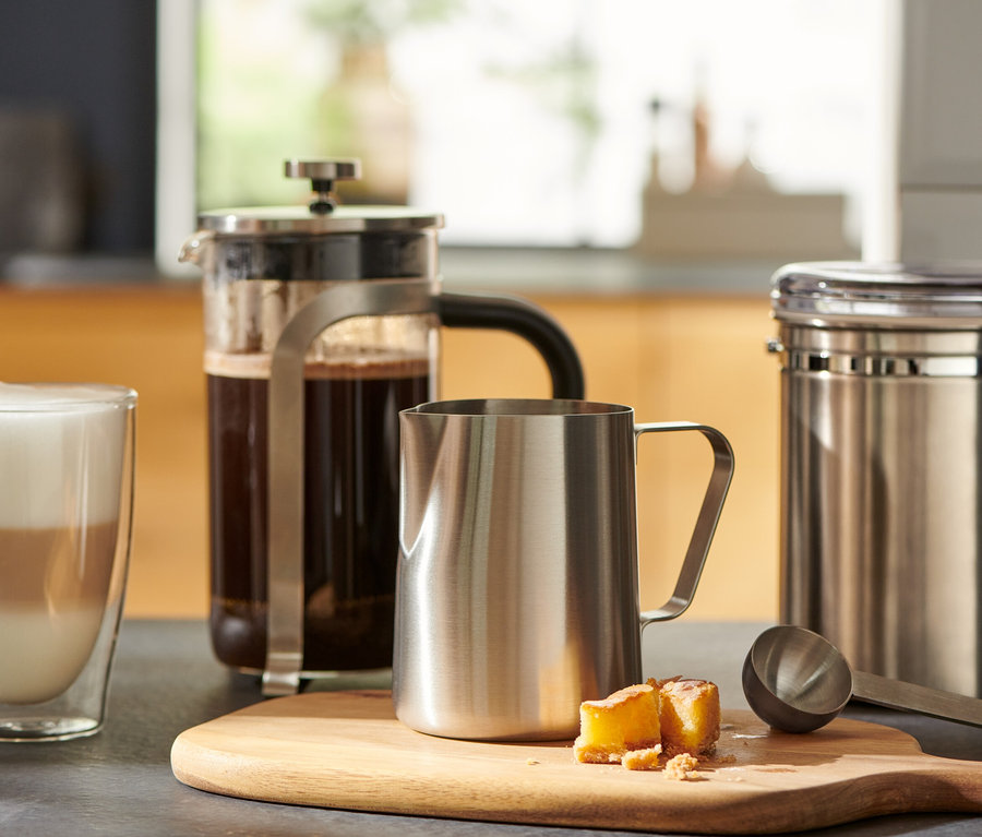 Un verre de café est posé à côté d'une presse française et d'un petit pichet à lait en acier inoxydable sur une planche à découper en bois.
