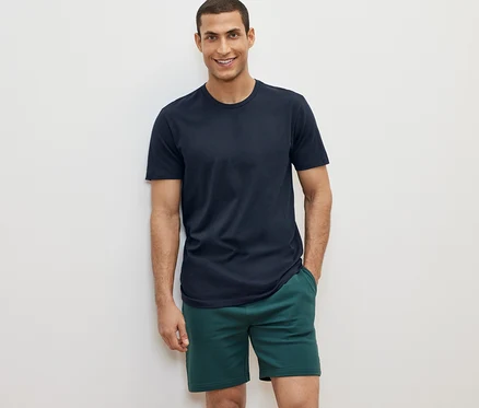 Un homme pose sur un fond blanc portant un des 2 t-shirts à encolure ronde et un short vert.