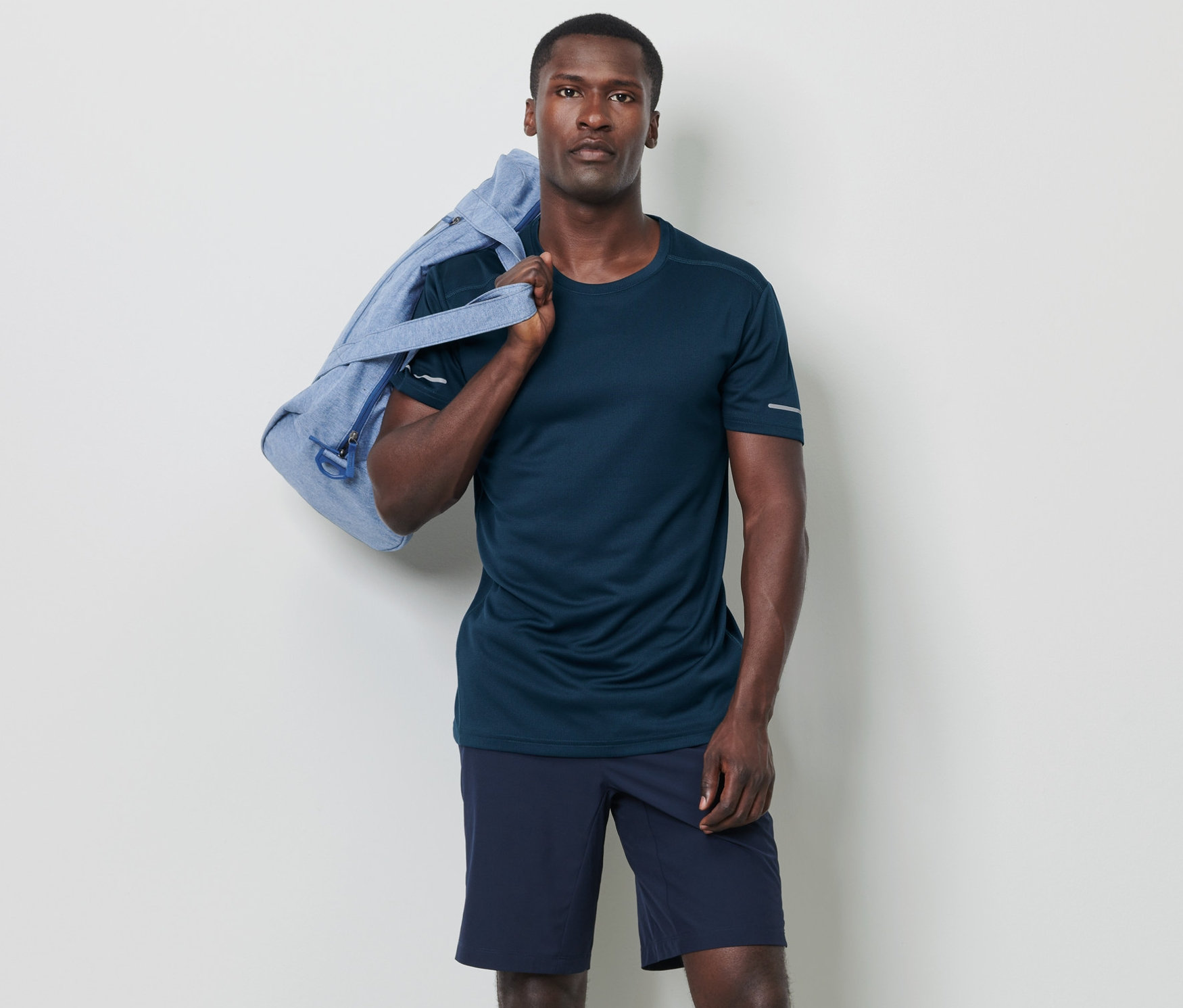 Un homme pose avec un sac bleu sur l'épaule et porte un t-shirt fonctionnel bleu foncé chiné.