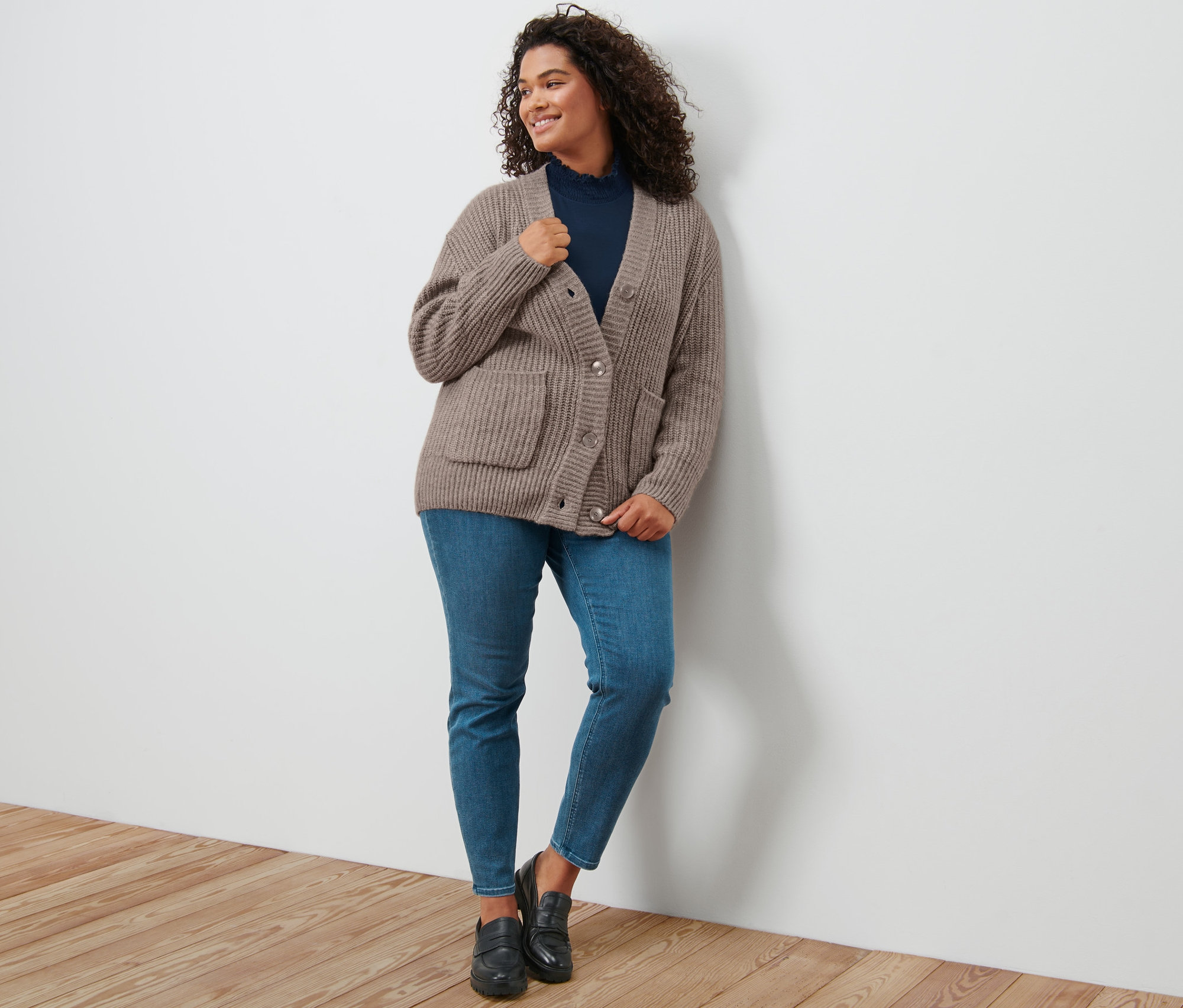 Une femme appuyée contre un mur porte un cardigan en grosse maille côtelée taupe, une blouse à col montant bleu foncé, un tregging «
Aucun
Aucun
Fit Nelly » et des loafers en cuir.