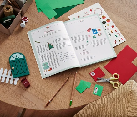Sur une table en bois sont posés un livre ouvert, des ciseaux, des crayons de couleur, des papiers colorés et des décorations, dont le livre « Hurra, ein Wichtel zieht ein! » (en allemand).