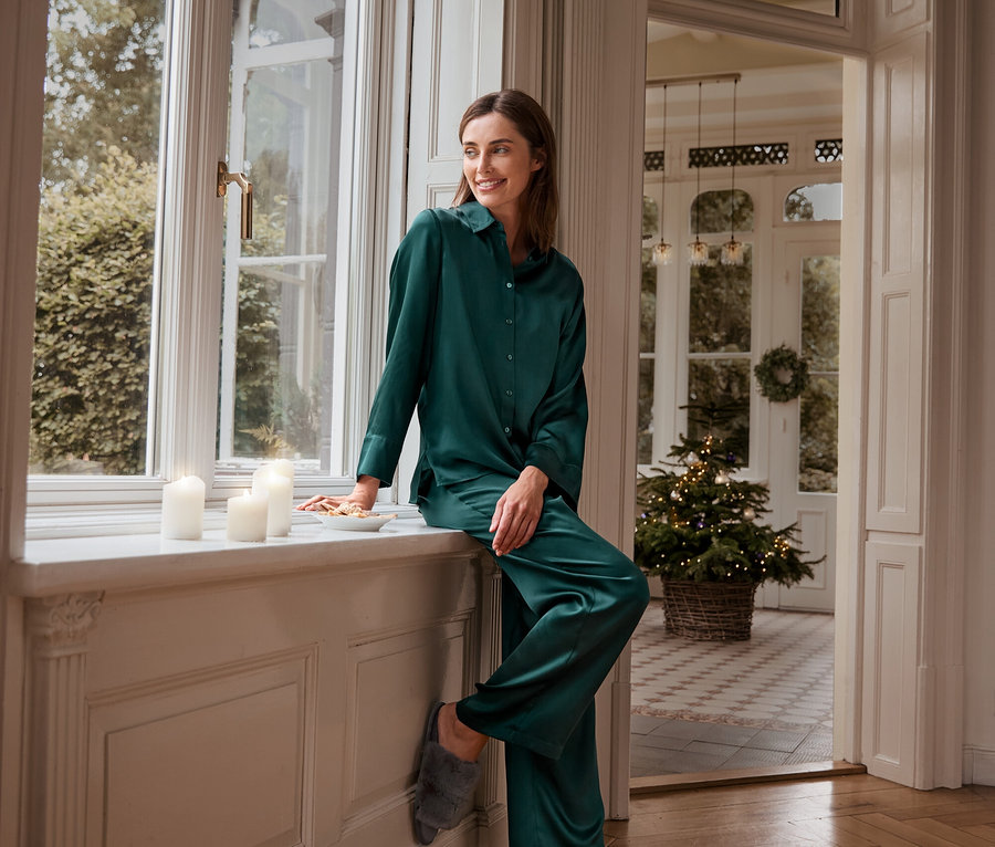 Une femme assise sur un rebord de fenêtre en pantalon et haut de pyjama en soie verte avec des chaussons en peluche.