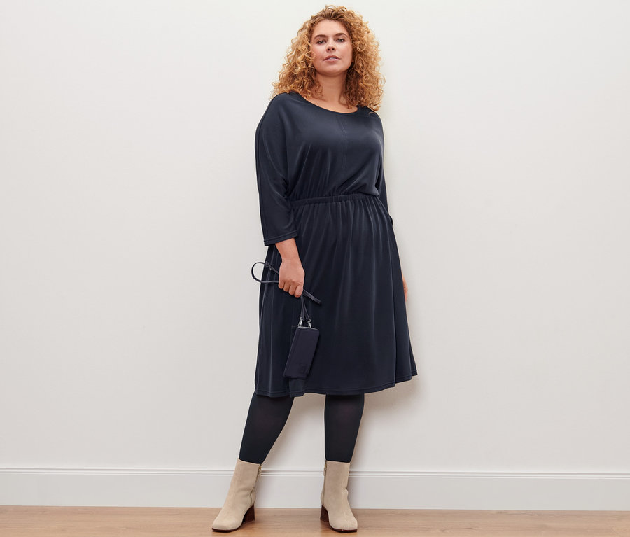 Une femme pose en robe en jersey piqué et tenant un mini sac. Elle porte 2 paires de collants fins.