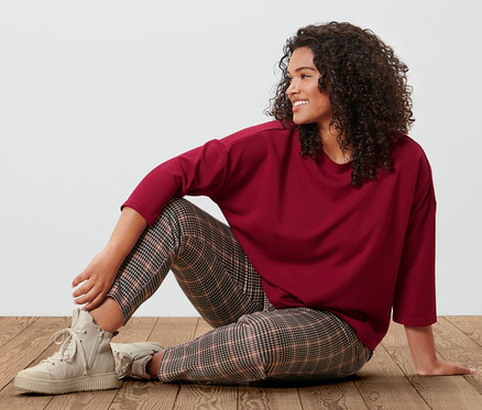 Femme assise sur un plancher en bois portant un sweat-shirt manches 3/4 rouge bordeaux et un jogpant à motif pied-de-poule.