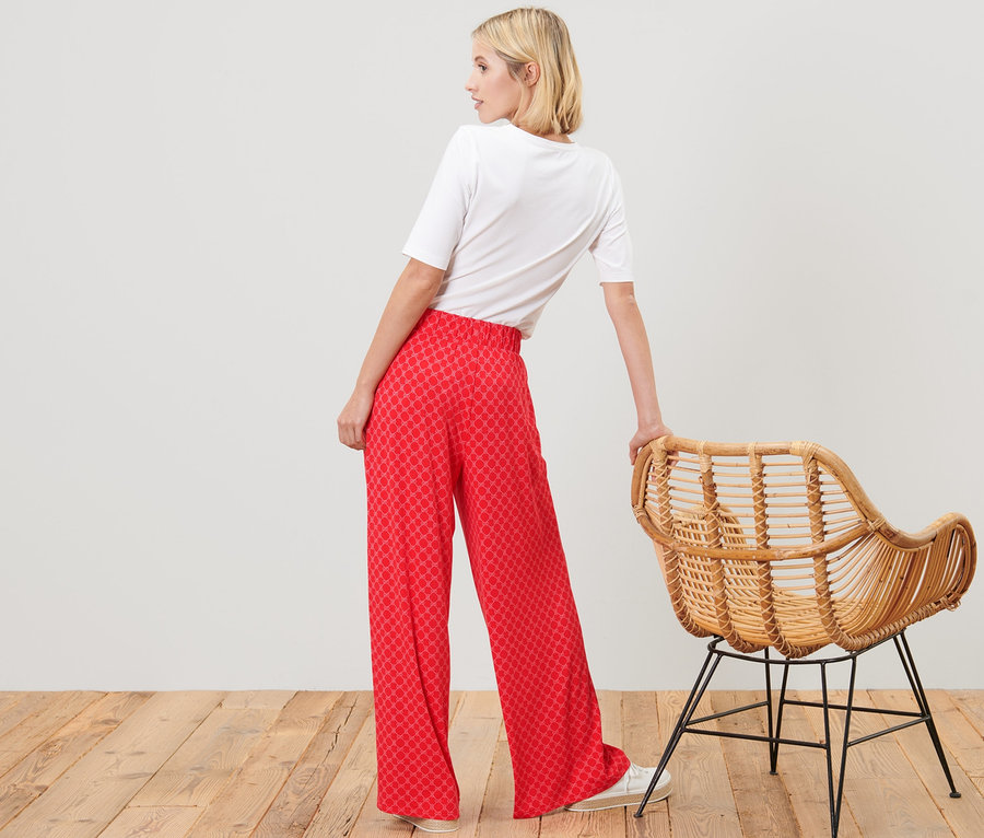 Une femme pose en pantalon palazzo imprimé rouge et un t-shirt blanc à côté d'une chaise en rotin.