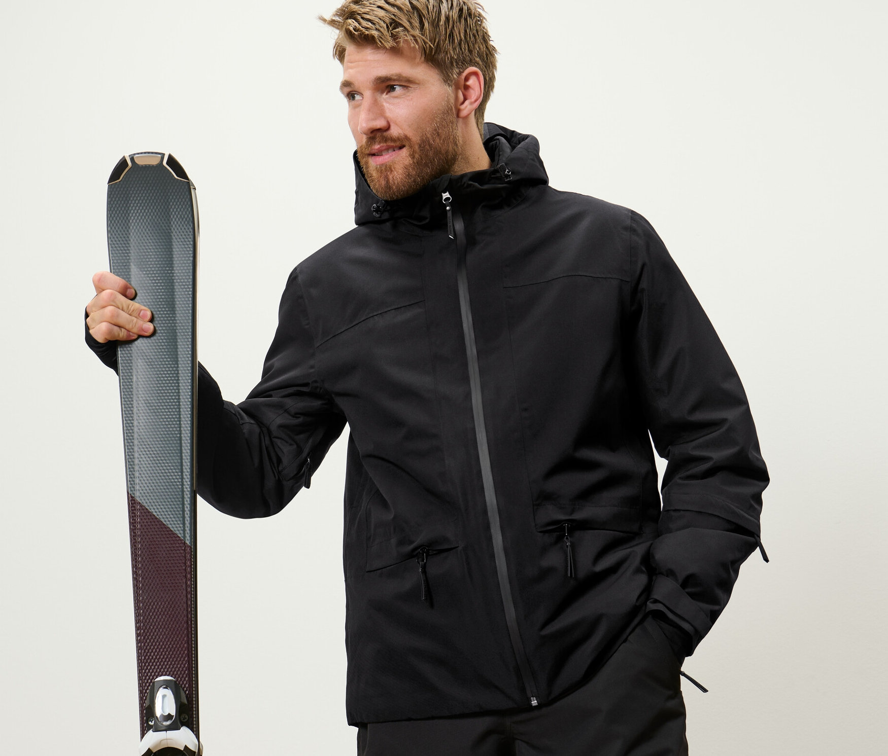 Un homme tient des skis. Il porte une veste de ski noire.