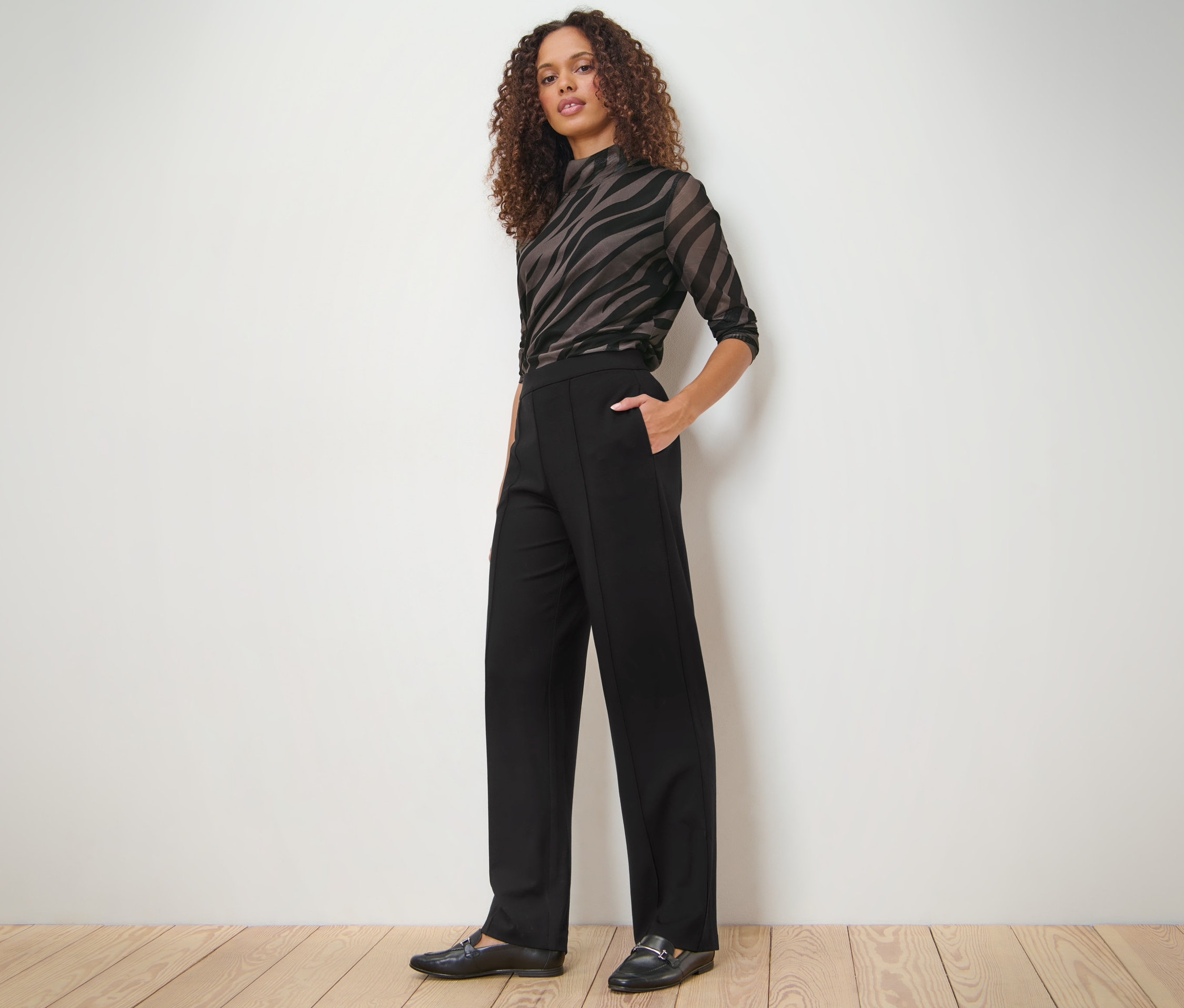 Une femme pose en pantalon en jersey noir avec passepoil, un haut zébré et des mocassins noirs.