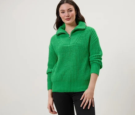 Une femme portant un pull camionneur en grosse maille vert avec un jean noir.
