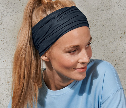 Une femme blonde aux cheveux attachés en queue de cheval porte un foulard multifonction et un haut bleu.