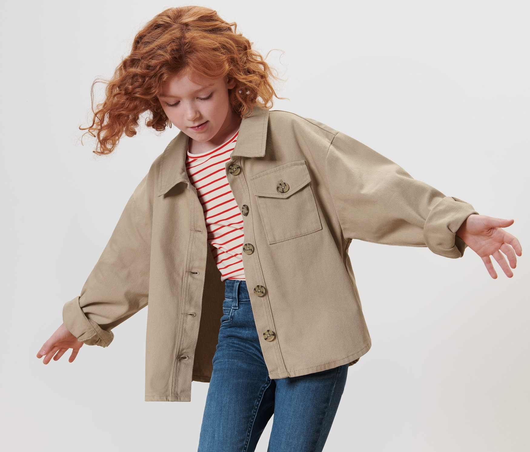 Une fille aux cheveux roux bouclés agite les bras et porte une veste en denim pour enfant.