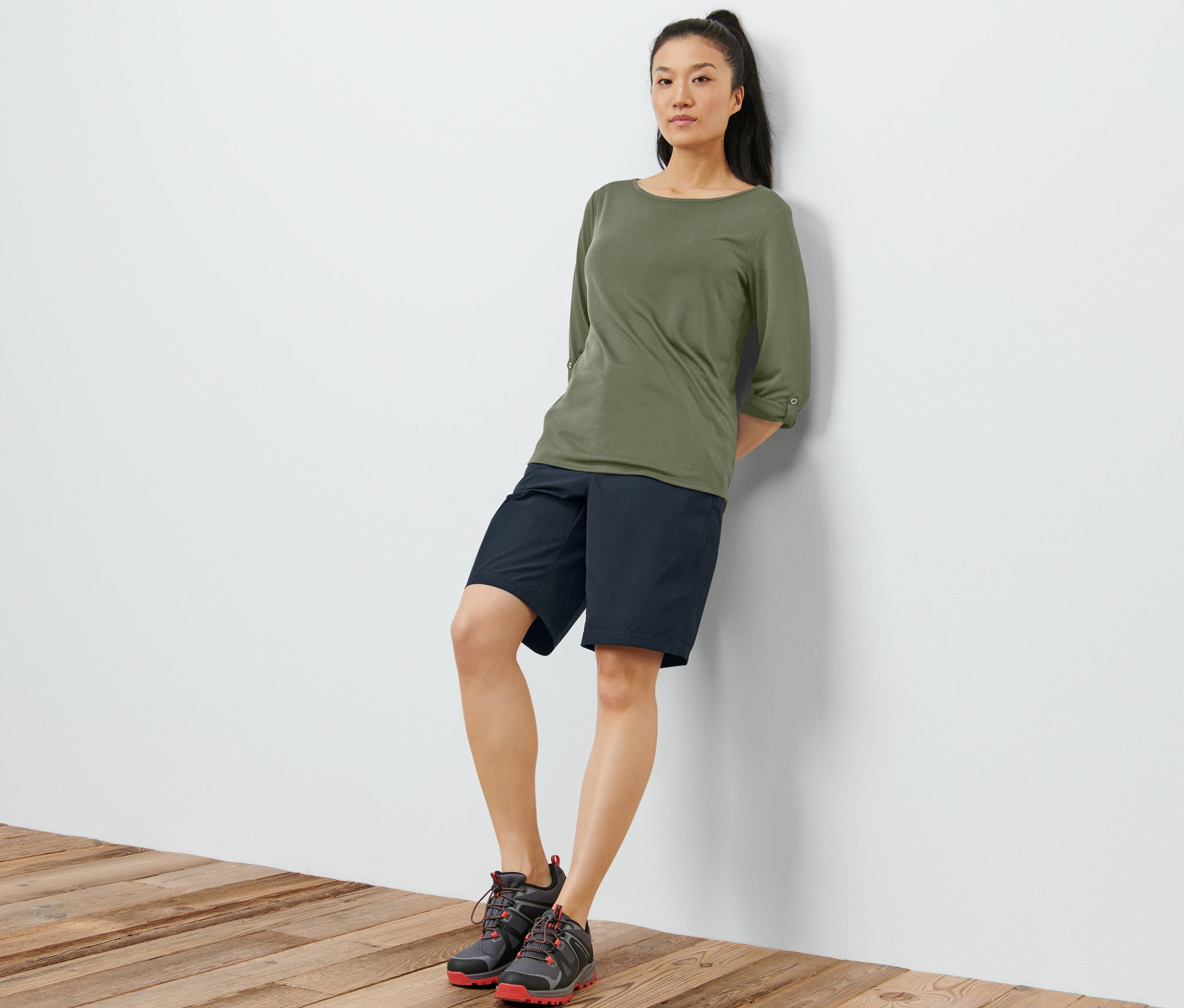 Une femme appuyée contre un mur blanc porte un t-shirt technique kaki à manches 3/4, un short noir et des chaussures de randonnée.