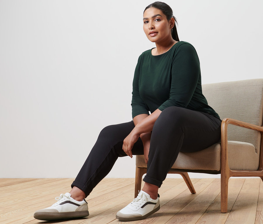 Une femme est assise dans un fauteuil et porte un haut manches 3/4, un pantalon de jogging et des baskets en cuir.