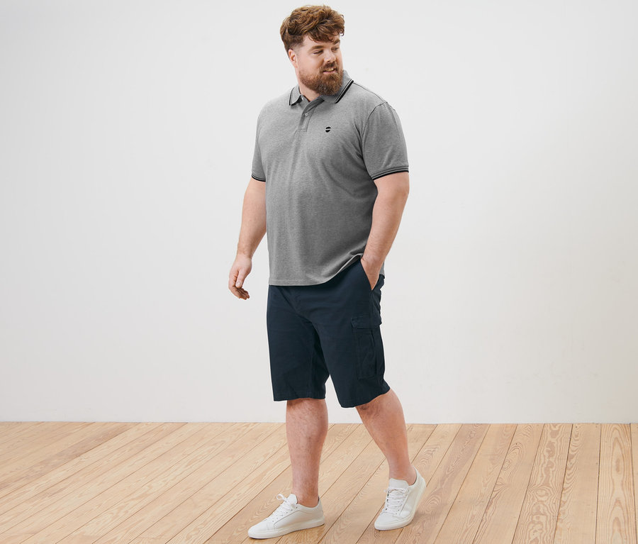 Un homme marche sur un plancher en bois et porte un polo en piqué gris et un short cargo bleu marine.