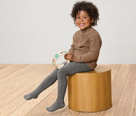 Un enfant souriant est assis sur un tabouret en bois et tient un tambourin. Il porte un pull marron et 2 collants thermiques pour enfant chinés gris.