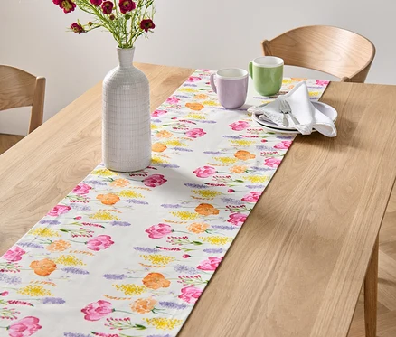 Sur une table en bois, un chemin de table blanc avec un imprimé floral. Un vase avec des fleurs et deux tasses sont posés sur le chemin de table.