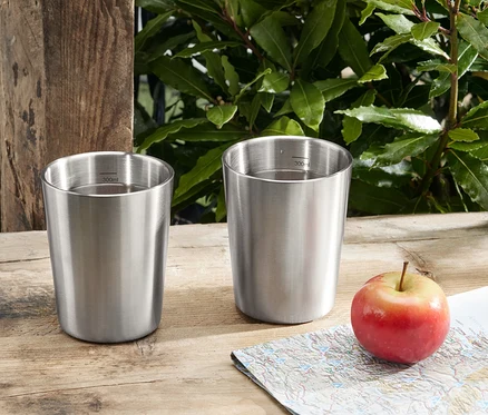 Deux gobelets de camping en acier inoxydable sont posés sur une table en bois à côté d'une pomme et d'une carte.