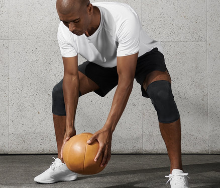 Un homme en T-shirt blanc et short noir tient un ballon de médecine et porte 2 genouillères de fitness.
