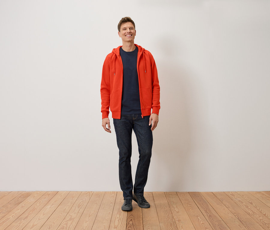 Un homme souriant debout portant un sweat à capuche orange, un t-shirt bleu marine et un jean.
