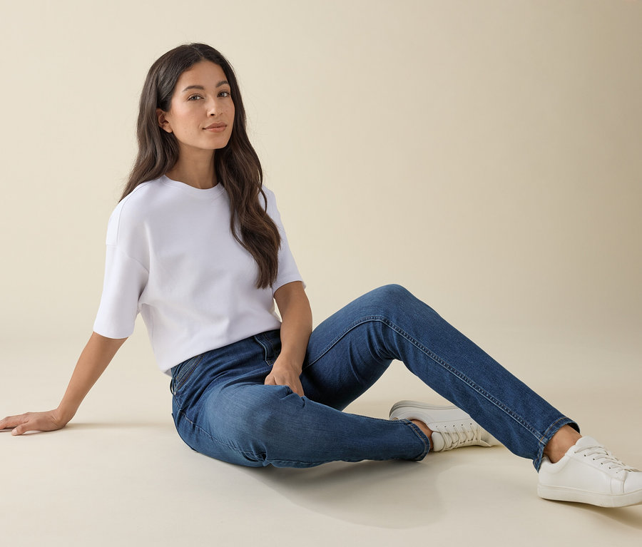 Femme assise par terre en jeans slim – Coupe « Emma », denim bleu moyen, t-shirt blanc et baskets.
