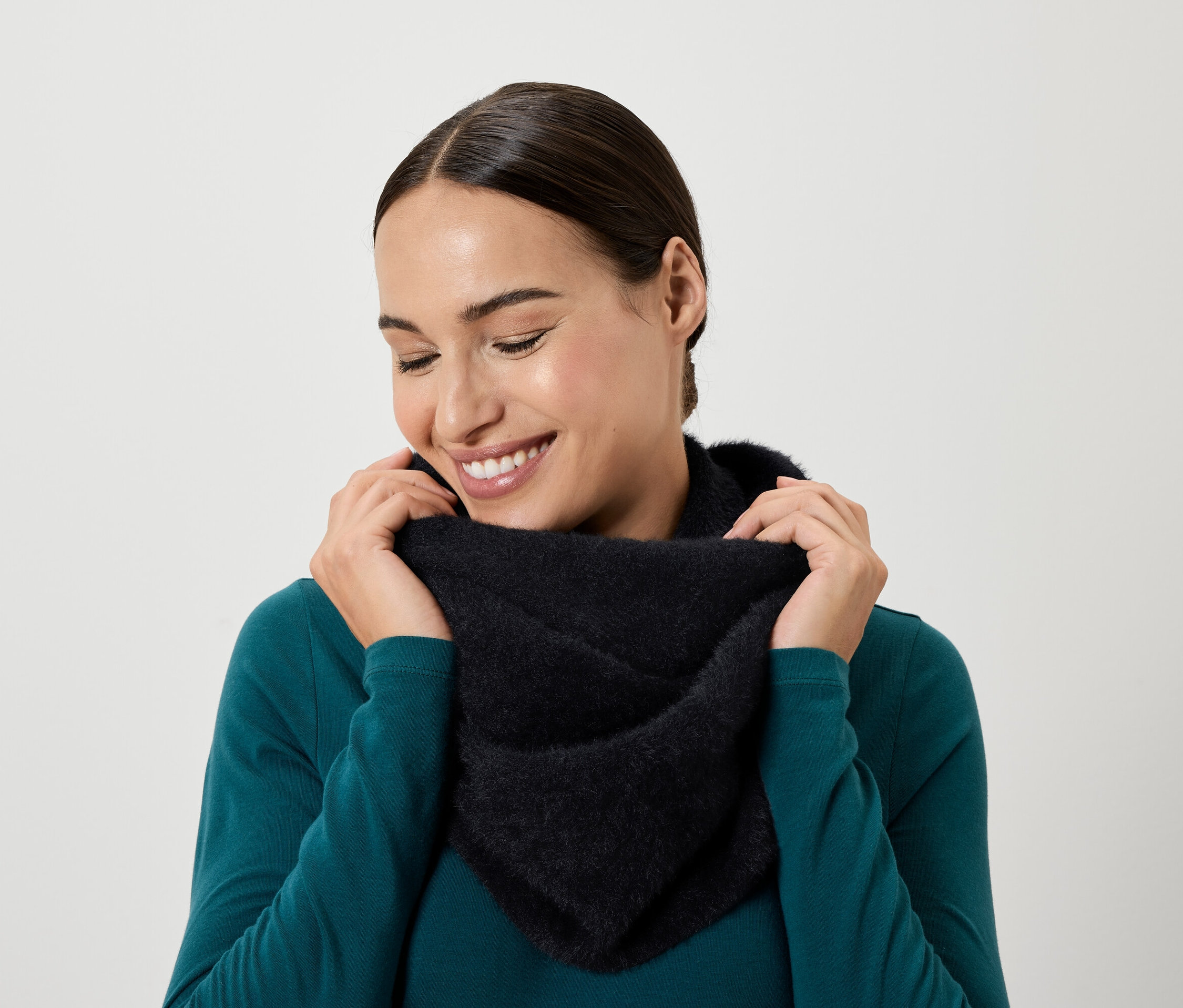 Une femme souriante regarde vers le bas et tient un foulard noir autour de son cou avec ses mains.